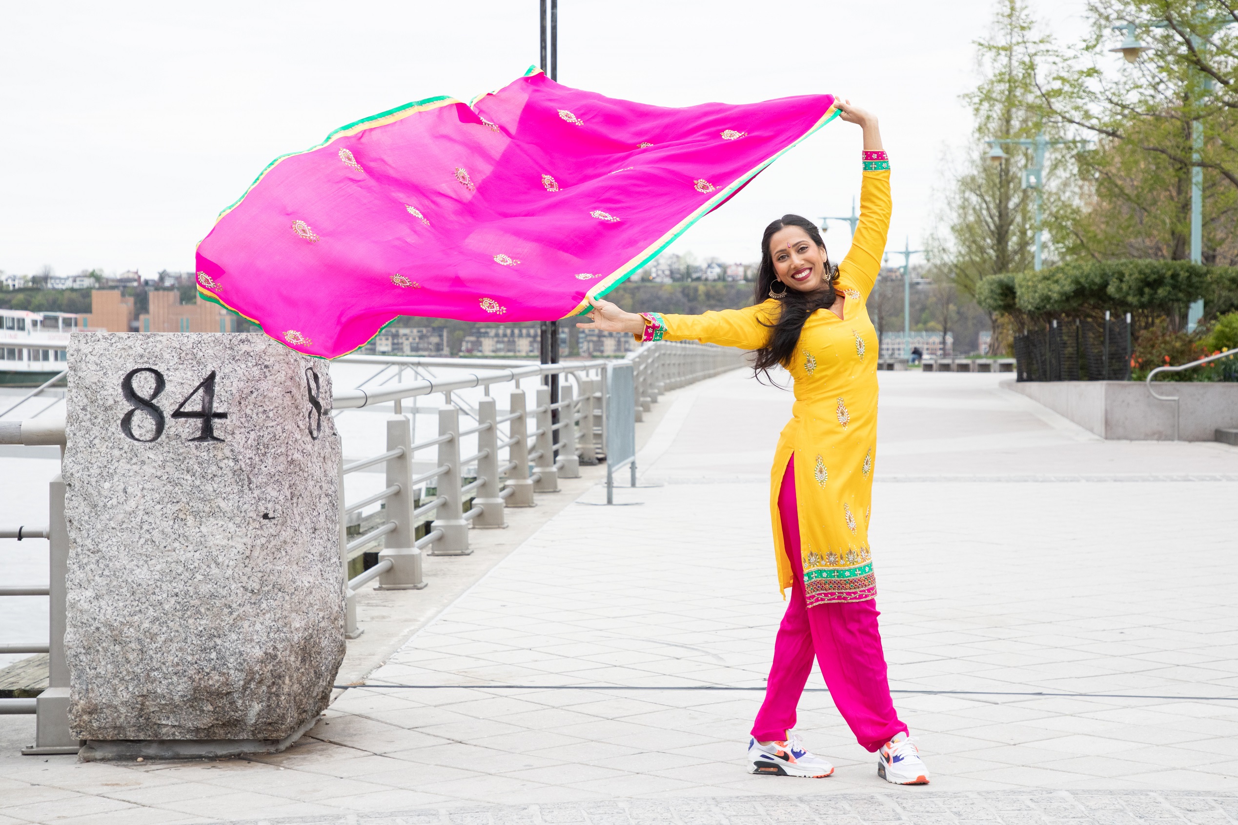 An image of a woman dressed in a bright yellow top and pink bottom holding a pink scarf that blows in the wind while smiling at the camera and standing on a boardwalk